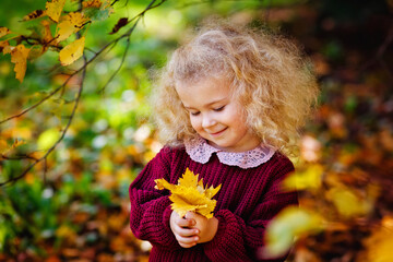 A small smiling blonde curly-haired girl in a burgundy sweater in an autumn park with a bouquet of...