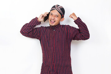 Fototapeta premium Happy joyful asian man with javanese traditional cloth lurik using mobile smartphone isolated on white background