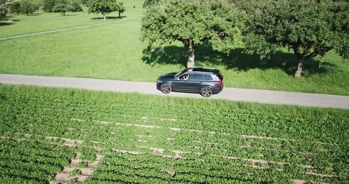 A Volvo xc 90 car drives on a small bumpy road in between fields, few trees, Swiss countryside, Vaud, drone aerial view