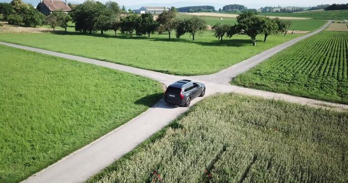 a volvo xc 90 car drives slowly before an intersection between two small roads before continuing straight ahead, swiss countryside and fields, aerial shot, drone