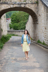 Fototapeta na wymiar Lifestyle portrait of young stylish woman with long brunette hair walking on the street in old town, wearing yellow dress and denim jacket