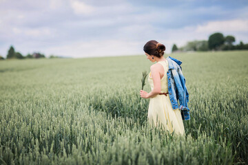 Lifestyle portrait of young stylish woman walking by wheat field and holding ears in his hands, wearing yellow dress