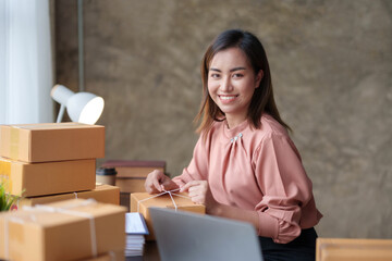 Happy young Asian business owner woman prepare parcel box and check online orders of product for deliver to customer on laptop computer. Shopping Online concept.