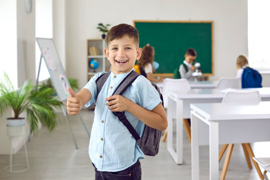 Funny little schoolboy showing thumbs up showing positive approval of great choice. Portrait of cheerful first grader with backpack on his shoulders standing in classroom and smiling at camera.