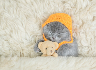 Fototapeta na wymiar Cozy tiny kitten wearing tiny warm hat sleeps with favorite toy bear under warm blanket on a bed at home. Top down view. Empty space for text