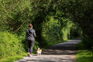 Slim athletic woman walking small dog on a leash in a park. Outdoor activity and healthy life style concept.