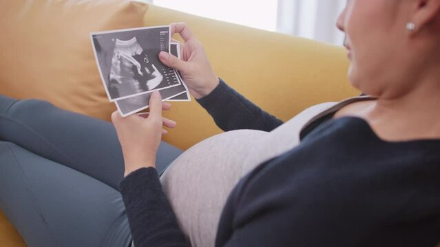 Happy Asian pregnant woman watching ultrasound image on sofa.