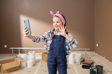 Little girl influencer in denim jumpsuit with headband stretches phone far away on hand waves hand...