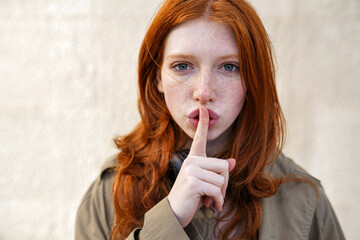 Hipster teen gen z redhead girl showing shh sign finger gesture asking to keep secret, be hush...