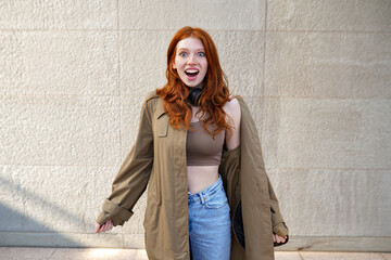 Happy excited teen redhead fashion girl standing on urban wall background looking at camera. Amazed...