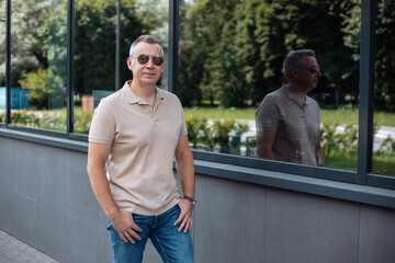 Portrait of senior brutal man in sunglasses walking with hands in pockets of jeans along modern office building, silhouette reflection in window. Urban lifestyle, outdoor recreation, summer leisure