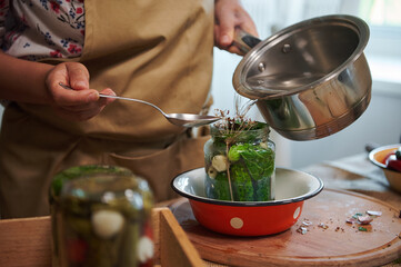 Close-up of a woman housewife in a chef's apron pouring boiling marinade over cucumbers in jars....