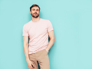 Portrait of handsome smiling stylish hipster lambersexual model. Sexy man dressed in pink T-shirt and trousers. Fashion male isolated on blue wall in studio