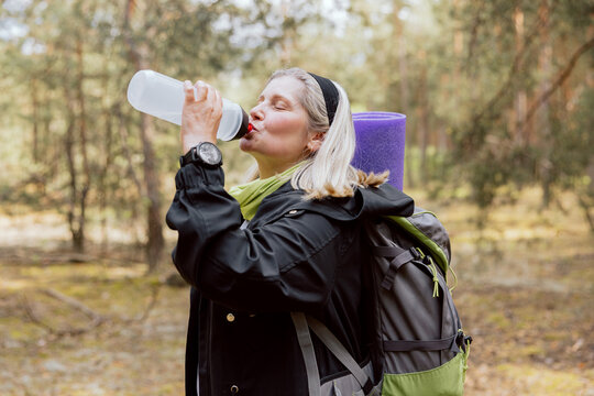 Close-up elderly woman with backpack and mat hikking in the woods drinking water having fun with her friends holding smartwatch in her hand relaxing.
