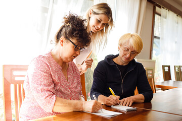 Young woman standing behind two elderly women, explaining business. Seniors sitting at desk, making notes in notepad.