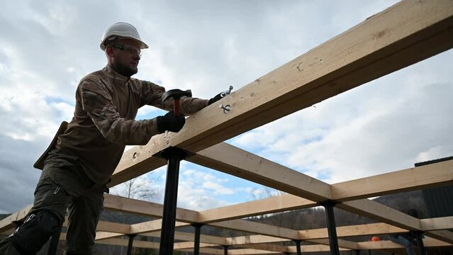 Man worker building wooden frame house on pile foundation. Carpenter hammering bolt into wooden joist, using hammer. Carpentry concept.