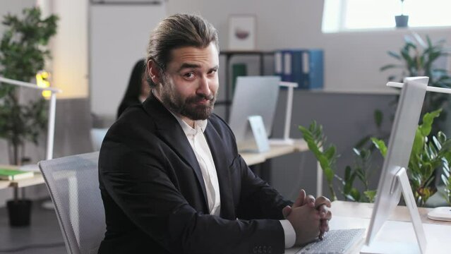 Handsome employee working on computer while sitting at business center room with female colleague on blur background. Young man raising eyebrow and having cunning look at camera.