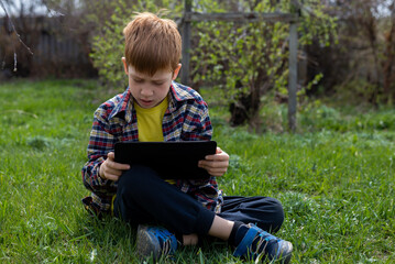 Happy child ginger boy playing on tablet or watching cartoons sitting on green grass in the backyard in the village, smiling