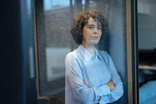 Thoughtful businesswoman standing with arms crossed seen through glass