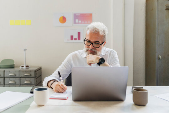 Senior businessman with hand on chin writing in notepad at office