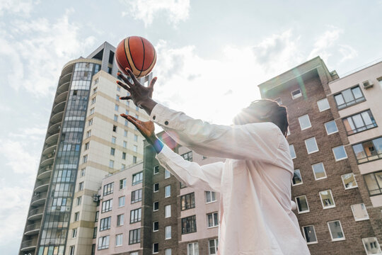 Young man throwing basketball in front of buildings on sunny day