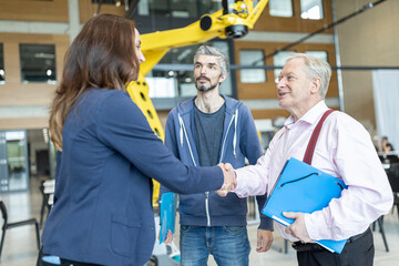 Senior technician shaking hands with female colleague in factory with industrial robots