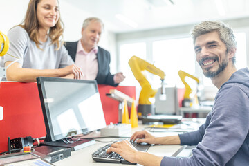 Happy colleagues watching smiling man working on PC in robotic factory