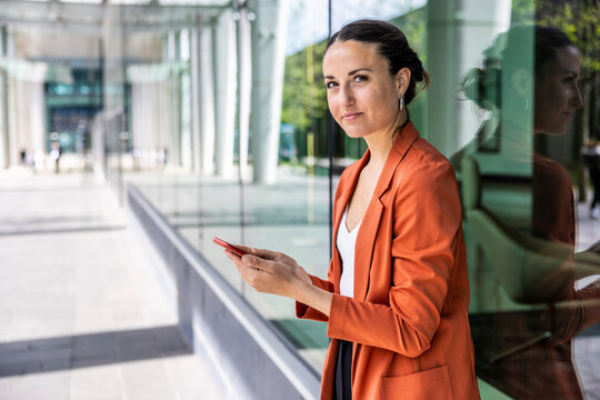 Businesswoman holding smart phone leaning on glass wall