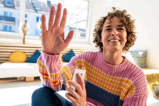 Smiling woman holding smart phone waving hand at home
