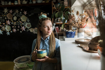Smiling craftsperson holding ceramic mug looking through window at workshop