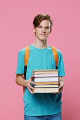 vertical photo of a student in a blue t-shirt with a stack of new books in his hands, standing on a pink background