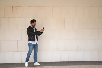 Handsome young man with beard, sunglasses, leather jacket, white shirt and jeans, next to a white wall, consulting his cell phone. Concept beauty, fashion, trend, smartphone, app, social networks.