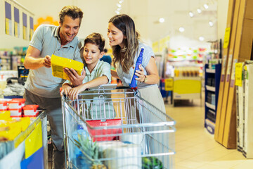 Happy family with child and shopping cart buying food at grocery store or supermarket - obrazy, fototapety, plakaty