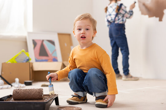 A 2-year-old child dressed in an orange blouse jeans and sneakers sits in middle of the room where repairs are being made. The boy holds paintbrush in hand and looks sweetly at the camera.