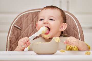 Happy toddler baby boy learns to eat porridge himself with a spoon while sitting in a child chair....