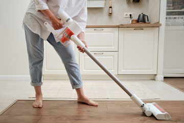 Woman with a wireless portable vacuum cleaner in the kitchen. Cleans the floor in an apartment with a vacuum cleaner with a battery - obrazy, fototapety, plakaty