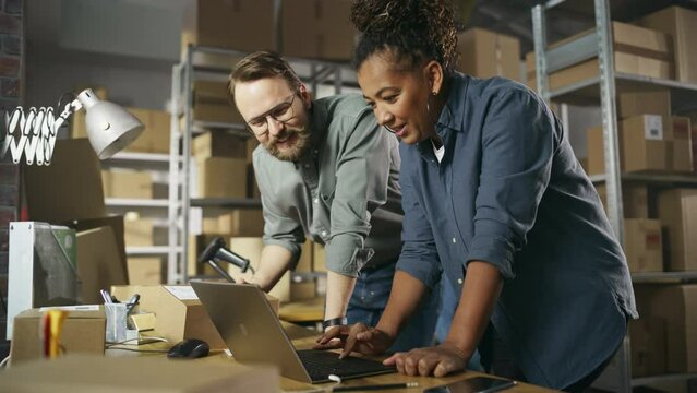 Happy Male and Female Working in Warehouse. Talking, Using Laptop Computer, Checking Retail Stock, Preparing Shipment. Successful Small Business Owners High Five Each Other.