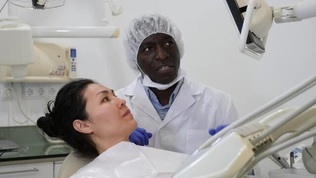 Asian woman patient looking at display during consultation in dental clinic. African-american man dentist standing beside.