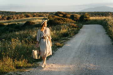 Portrait of a stylish woman walking with a straw bag through the field at sunset