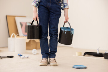 Fototapeta premium The child dressed in jeans and checked shirt holds cans of paint in hands. The girl's face is not visible, only new ones. In background are paintings, paint rollers, paint and other things for repair.