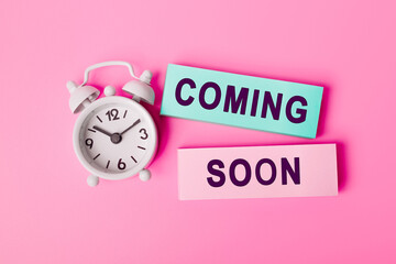 White alarm clock isolated on vivid pink background with text COMING SOON, written on paper notes....
