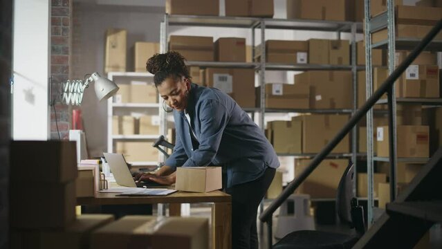 Warehouse Female Inventory Manager Using Laptop Computer, Preparing a Small Parcel for Postage. Black Multiethnic Small Business Owner Working in Storeroom, Preparing Order for Client.