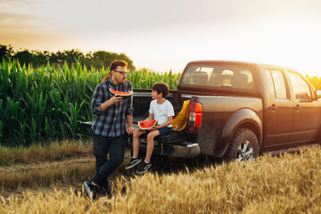 father and son sitting on trunk of truck and talking. they are outdoor in field eating watermelon