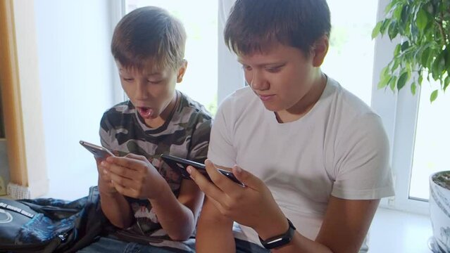 Two boys play computer games at home on their smartphones, watch videos, rejoice and laugh. Two brothers watch interesting videos together on tablet or smartphone. They laugh and indulge. Sit on table