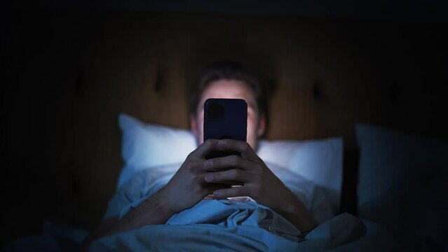 Caucasian Man Uses Smartphone in Bed at Home at Night. Handsome Guy Browsing Social Media, Reading News, Doing Online Shopping, Chatting with Friends Late at Night. Focus on Hand Holding Mobile Phone