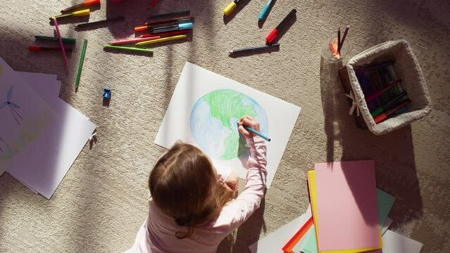 Top View: Little Girl Drawing Our Beautiful Green Planet Earth. Child Having Fun at Home on the Floor, Imagining Our Planet as a Happy Place with Clean, Sustainable Living. Cozy Sunny Day. Zoom out