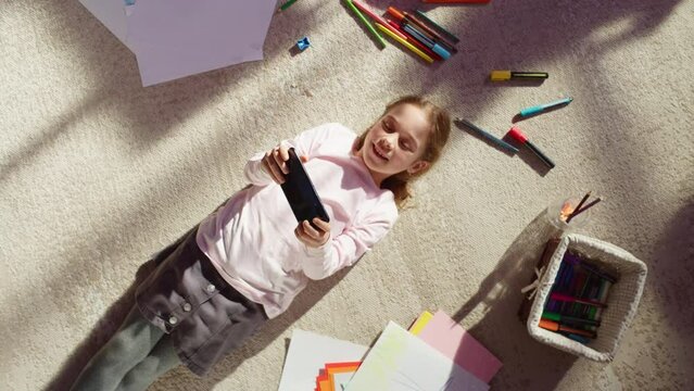 Top View Home: Happy Little Girl Using Smartphone While Lying on the Floor. Joyful Sunny Day and Clever Smiling Child Playing Smart Video Games, Using e-Learning Apps on Mobile Phone. Zoom Out Shot