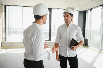 A front view of two smart architects with white helmets reviewing blueprints at a construction site on a bright sunny day