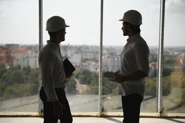 A front view of two smart architects with white helmets reviewing blueprints at a construction site on a bright sunny day