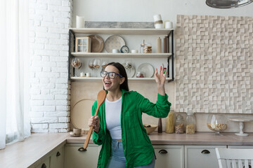Young beautiful woman in glasses and green shirt dancing and singing at home in the kitchen. The...